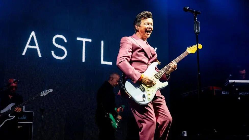 Rick Astley wearing a pink suit, playing guitar on stage