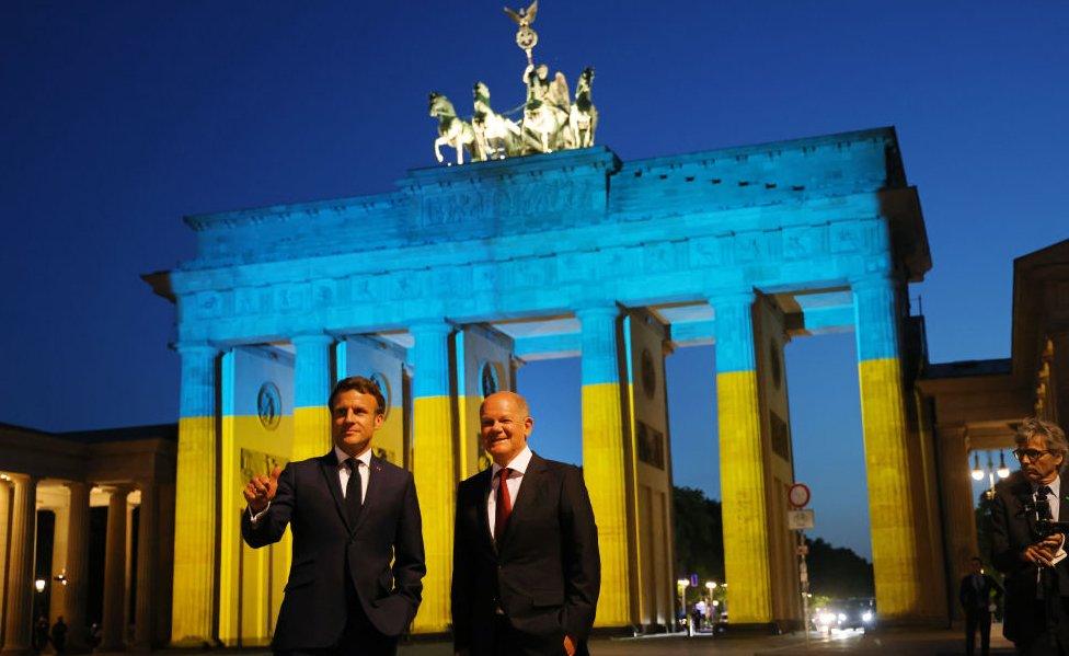 German Chancellor Olaf Scholz (R) and French President Emmanuel Macron stand in front of the Brandenburg Gate that is illuminated in the colours of the Ukrainian flag following talks at the Chancellery on May 09, 2022 in Berlin, Germany