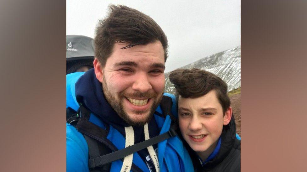 Michael Hall and Joshua Hall are huddled together grimacing at the camera with a snowy mountainside in the background.