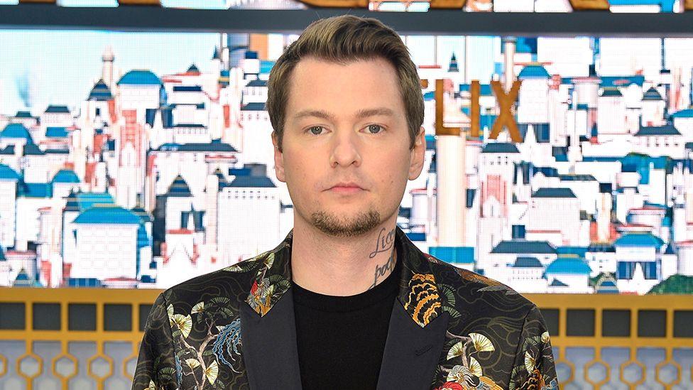 A man with neatly cropped hair stands in front of a large picture of a town made up of tall, bright white buildings. It looks Meditteranean. He has handwriting tattooed on his neck and wears a black blazer with a traditional Japanese pattern featuring cranes and paper fans.