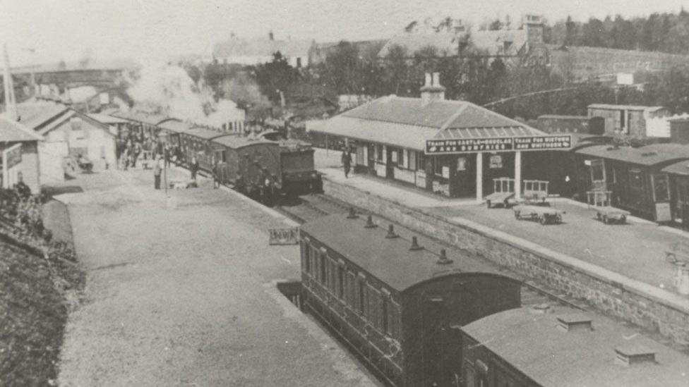 An old station on the Dumfries to Stranraer line in black and white with a train in the station and passengers milling about