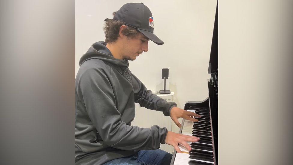 Brad Kella, wearing a baseball cap, tracksuit top and jeans playing a piano