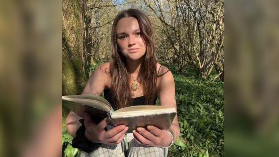 Kit Birks sitting smiling at the camera has long brown hair and brown eyes, she is holding a book.