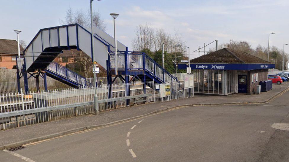 A general view of the train station, with a small ticket office can be seen on the right next to a car park. There is a a pedestrian bridge over the tracks joining two platforms. 
