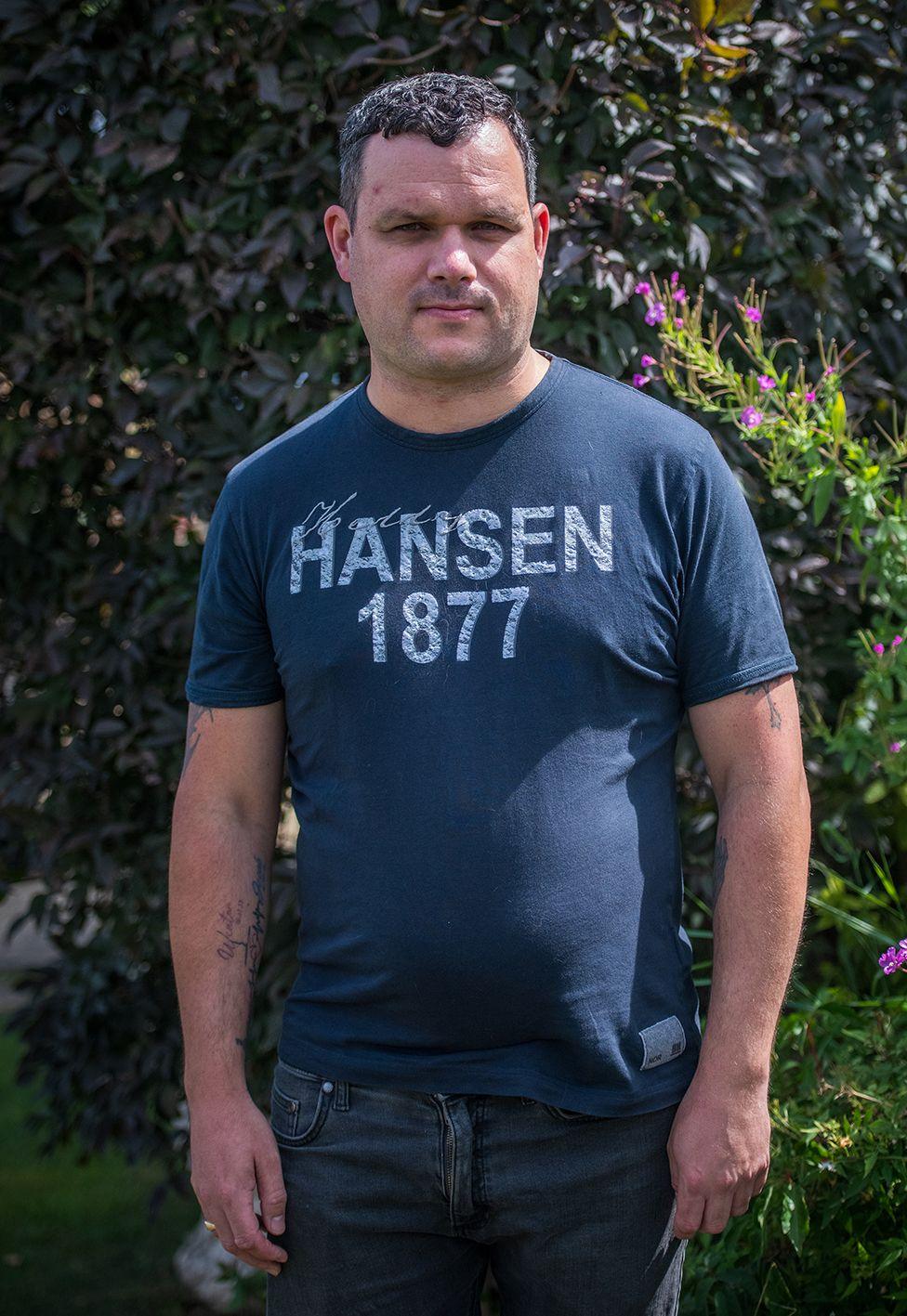 Ben, the father of Alice's eldest daughter Delsie. He is wearing a blue jeans and a t-shirt with Hansen 1877 logo on it. He as short dark brown hair.