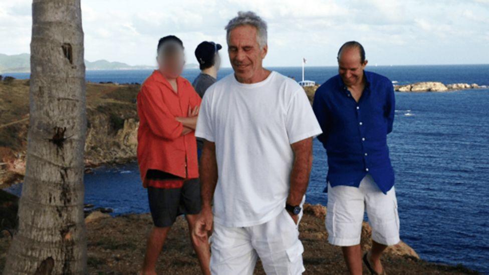 Several people stand outdoors beside a coastline overlooking the sea. In the foreground, Jeffrey Epstein in a white T-shirt and Howard Lutnick in a blue button-down shirt are visible, with rocky outcrops and deep blue water in the background under a cloudy sky. Two other people, blurred, stand to the left.