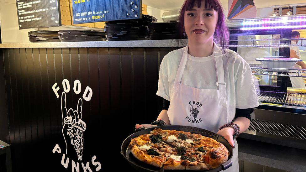 A young woman with long purple hair holds out a pizza on a plate in front of a food counter and kitchen