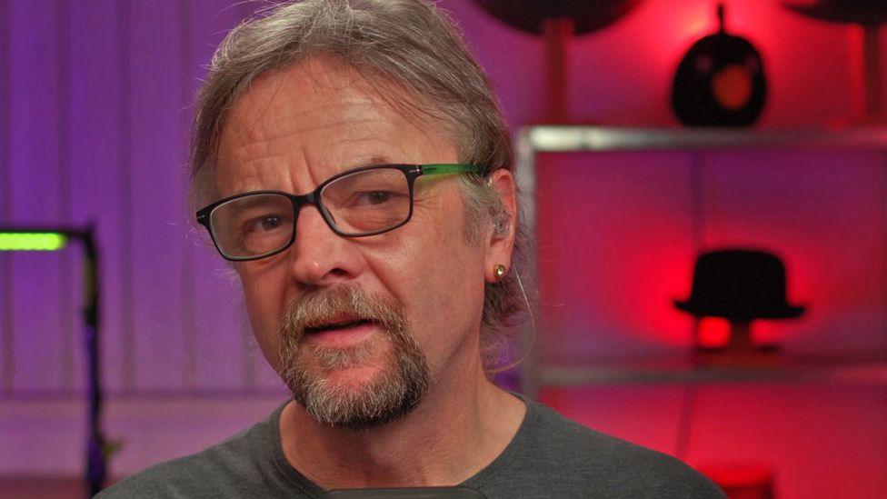 Paul Duckett wearing glasses and a grey T-shirt. The wall behind him is lit purple and red