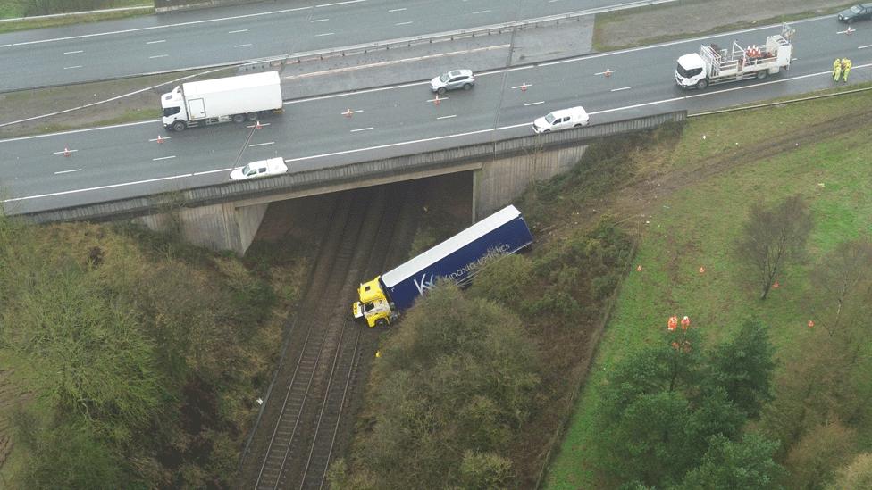 A drone shot of a  yellow and blue lorry at the bottom of a grassy embankment on railway lines that run below a road bridge, with several white vans and other vehicles parked on the road above the line.