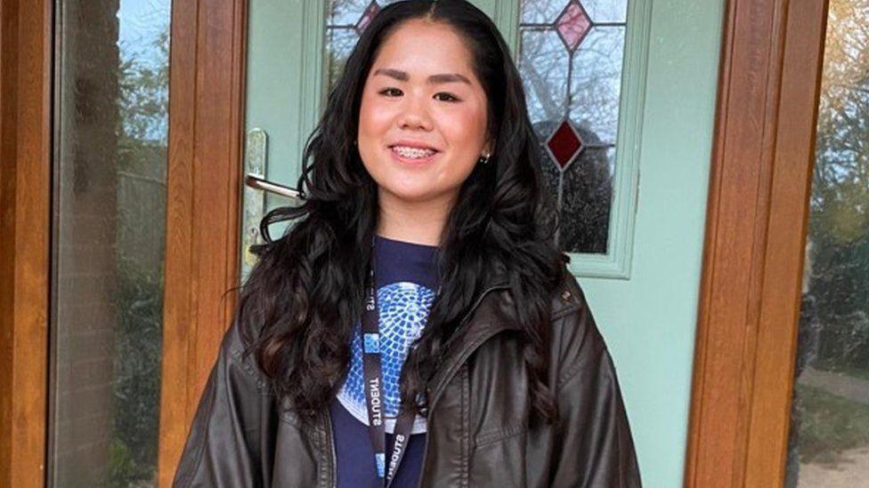 Teenage girls with long dark hair smiling at the camera in front of a door -she wears a black leather jacket and has a lanyard around her neck.
