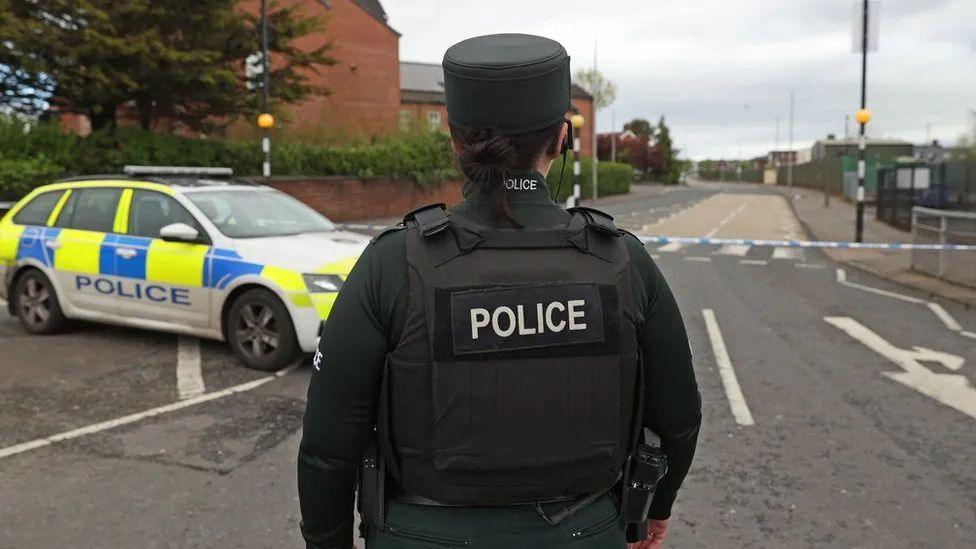 A PSNI officer standing with her back to the camera. She is wearing police uniform and has her brown hair up in a bun. She is standing in front of a parked police car and closed off road. 