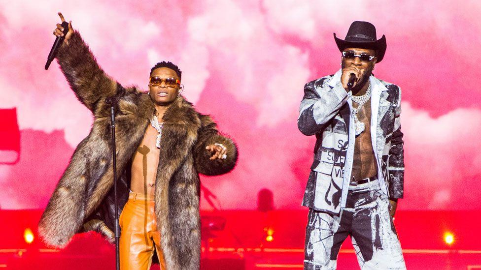 Wizkid in orange trousers and fur coat and Burna Boy in a black and white suit with black Stetson sing on stage together