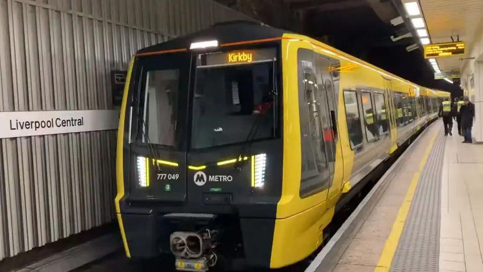A yellow and black Merseyrail train at a platform at Liverpool Central station. The destination Kirkby is displayed in yellow at the front of the train which carries the Metro logo.