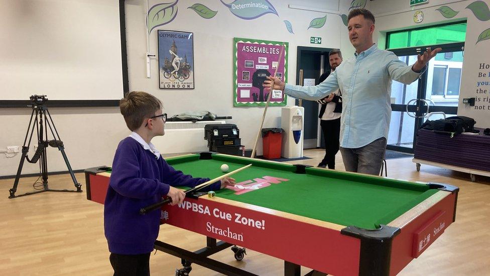 Boy in school jumper holds a snooker cue on a table while Kyren Wilson stands opposite with arms outstretched.