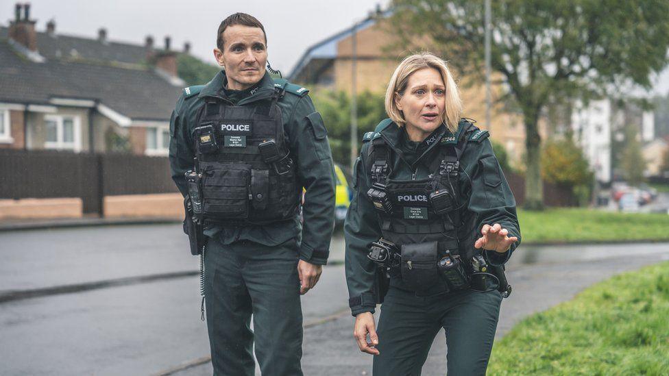 Actors Martin and Siân are dressed in PSNI uniforms during a scene the TV show blue lights. They are filming in a residential area with grass and road in the background.