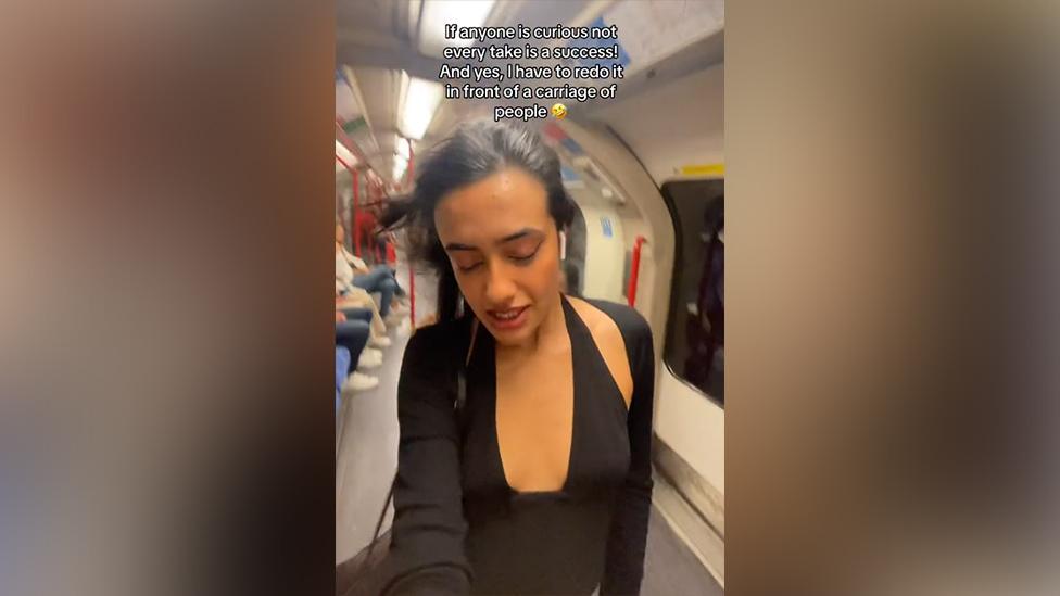 A still from a video of young Asian woman dancing on a Tube carriage. She's got her eyes closed, long black hair blowing behind her in the breeze from the Tube window. She's wearing a black cardigan over a black halter top. Behind her other passengers are sitting down - it looks quite busy. A caption on the still reads: "If anyone is curious not every take is a success! And yes, I have to redo it in front of a carriage of people [cry/laugh emoji]".