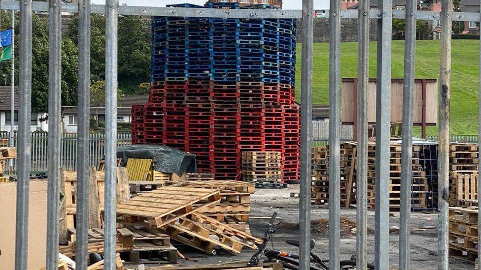 pallets and other materials for a bonfire seen through a hole in iron railings
