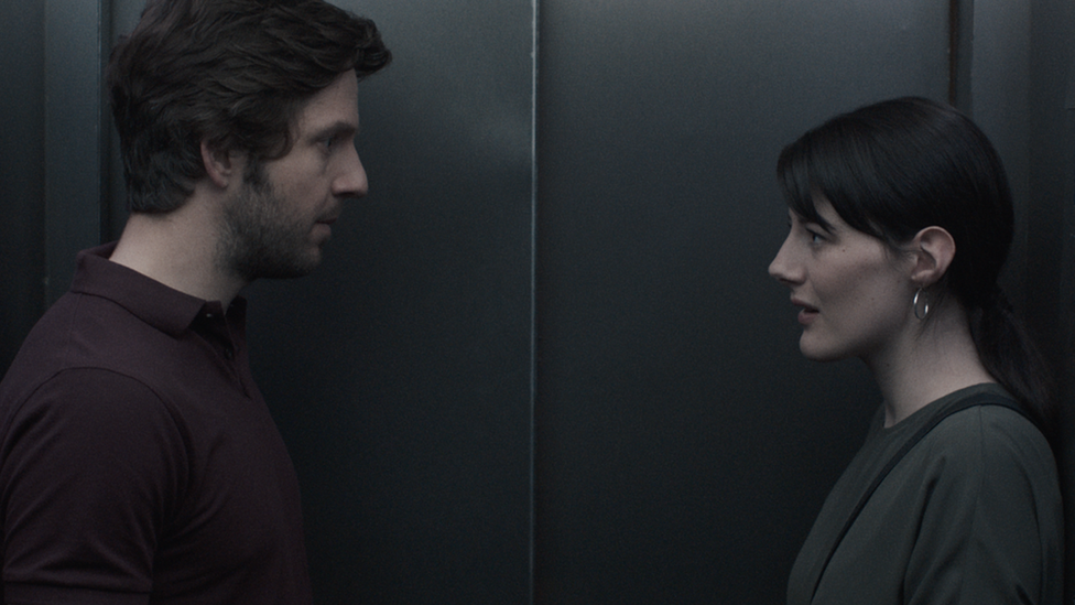 A still from a film. A man and woman stare at one another inside a lift.