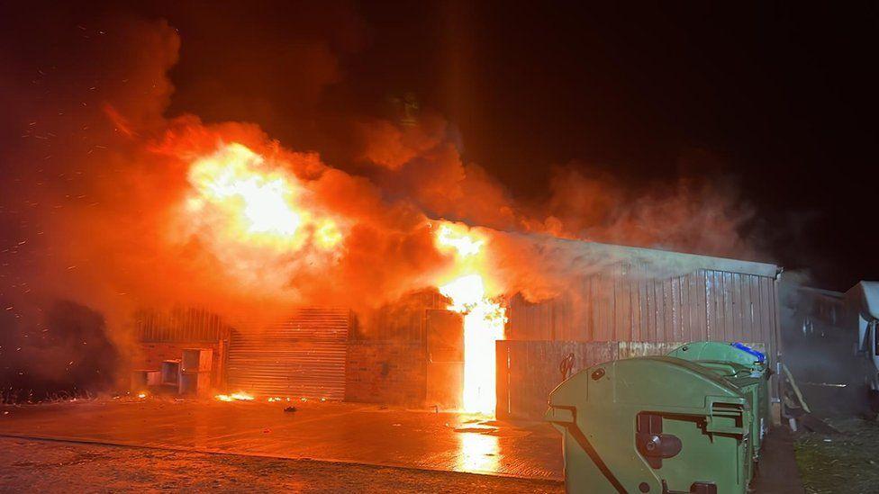 Large green bins lined up on the right in front of corrogated large building inside is glowing with fire and smoke is pouring out.