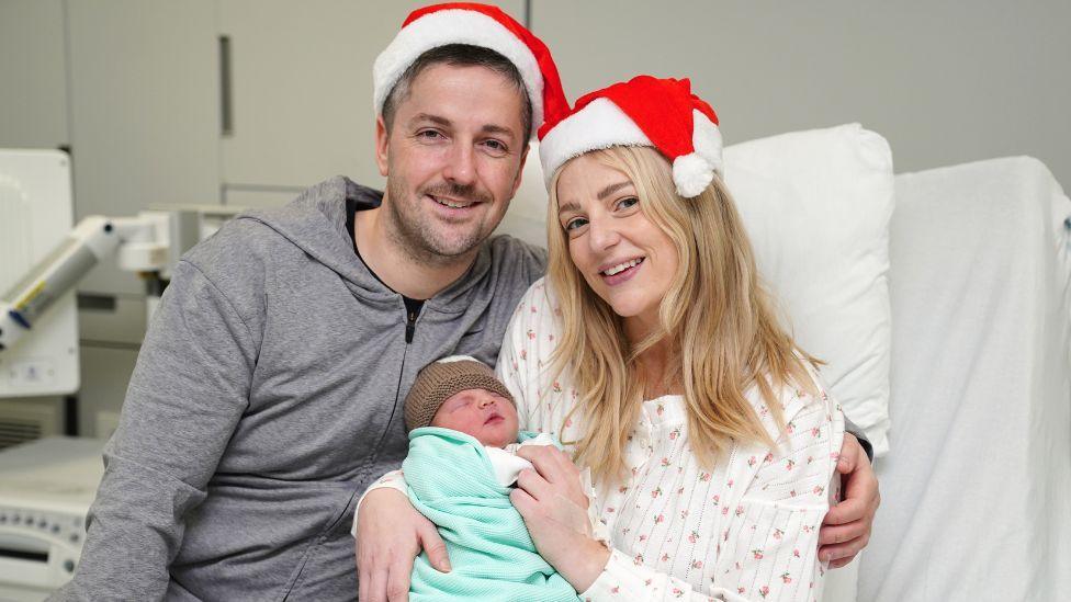 A smiling couple wearing Santa hats hold their newborn baby, wrapped in a blue blanket, while sitting in a hospital bed