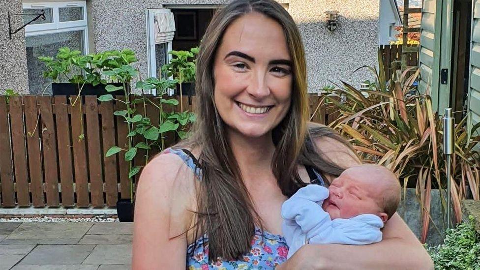 A woman with long brown hair smiling at the camera. She is wearing a blue flowery top and holding a new-born baby.