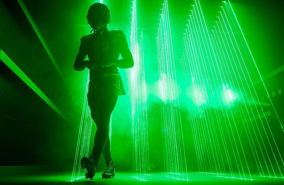 Silhouette of an American football player walking through bright green laser lights and smoke in a dark tunnel.