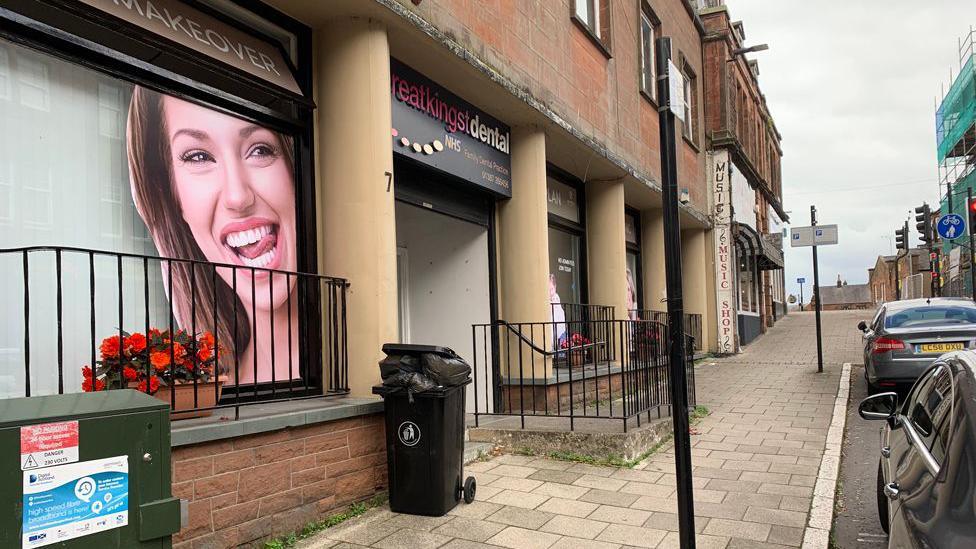 A dental practice with a picture of a smiling woman on its window, a bin out front and cars parked nearby