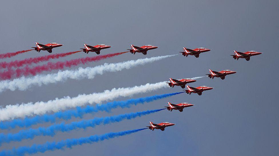 The nine aircraft of the modern Red Arrows flying past in a diamond formation trailing red, white and blue smoke