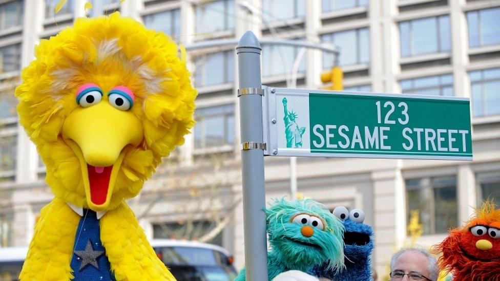 Big Bird and other Sesame Street puppet charactors pose next to temporarty street sign.