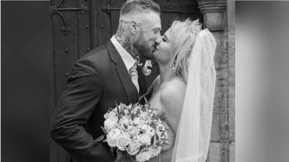 Aaron and Samantha Morris on their wedding day. It is a black and white photo and they are kissing while smiling outside a church door. The bride has bare shoulders, a tiara, white long veil and is holding a bouquet of roses.