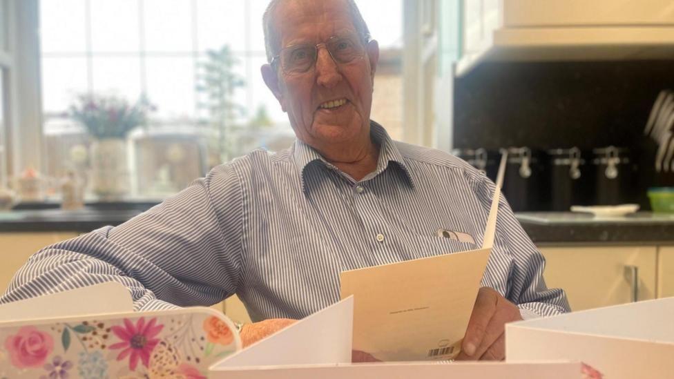 A man sat in a kitchen wearing a blue and white shirt holding gift cards in his hands.