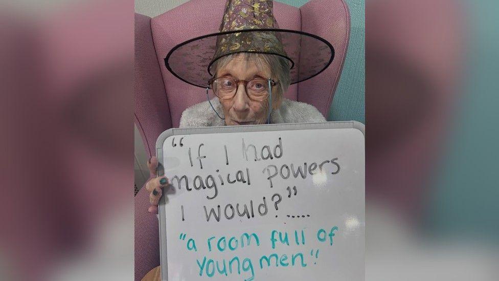 An elderly woman sitting in a pink chair wearing glasses and a witches hat holding up a sign which reads "if I had magical powers I would?" with the answer "a room full of young men".