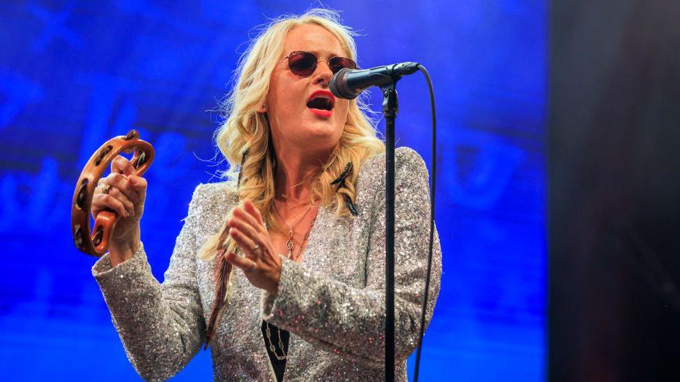 Elles Bailey has long blonde hair, and wears sunglasses and a glittering silver blazer. She is singing into a microphone on a stand, and holding a tambourine. 