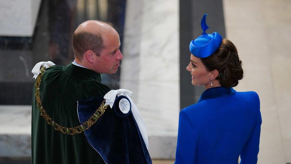 the Duke and Duchess, photographed from behind them at a high angle with the Duke wearing regalia and the Duchess in a blue outfit and fascinator