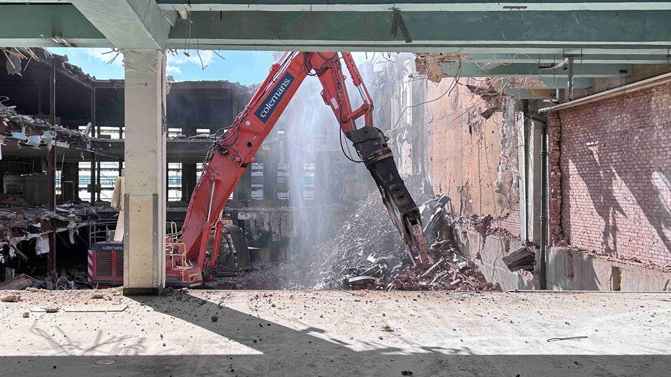 An orange excavator with a large jointed arm at a right angle as seen from a part-demolished M&S building in Northampton. The arm is in the middle and its grabber is reaching towards a pile of rubble. Beyond it is the rest of the three-storey part-demolished building which is open to the elements.