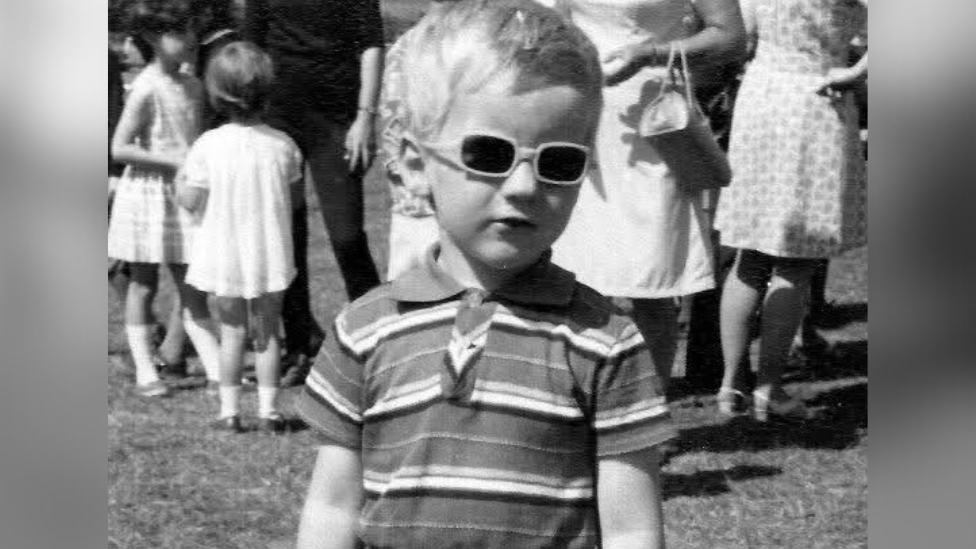The Image shows a black and white picture of a young boy wearing sunglasses and a striped shirt. Other children and woman are gathering in the back of the image. 