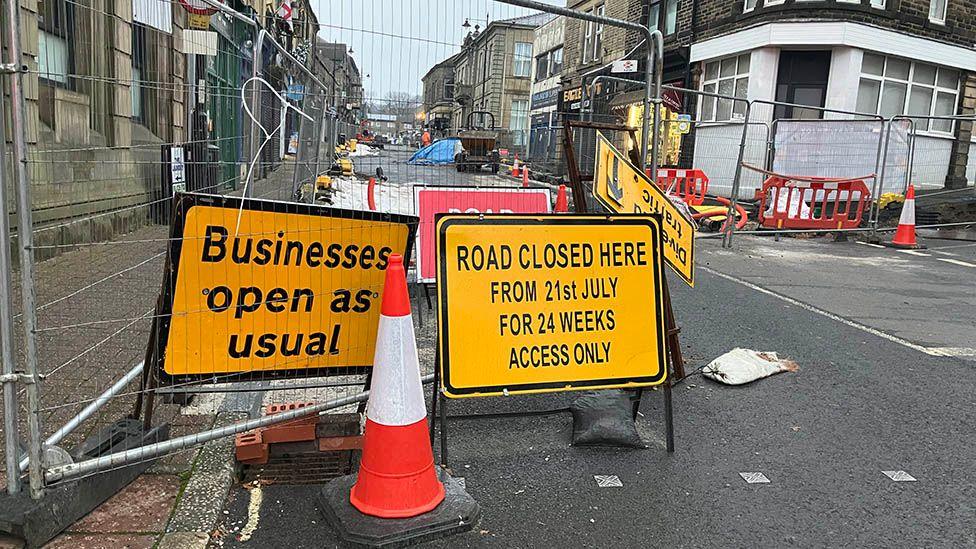 The street blocked off with metal fancing with yellow and black signs across the end saying "Businesses open as usual" and "Road closed"