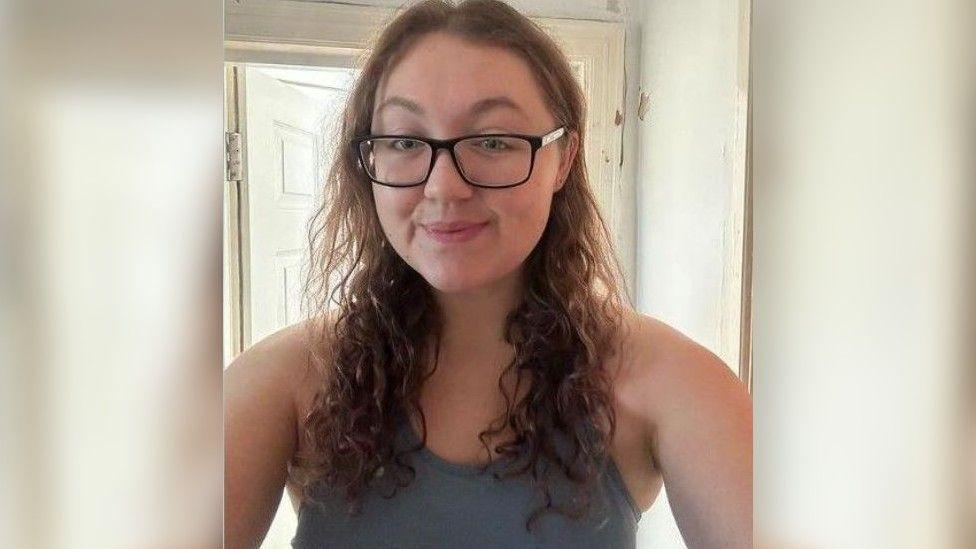 Gemma Peters, a young women with brown curly hair and black glasses wearing a grey top. She is smiling at the camera for a selfie. 