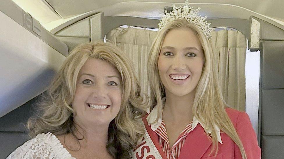 Close up of Vicky and Tabitha Bennett on an aeroplane. Both are blonde and smiling, Vicky's hair is wavy and Tabitha's is straight and she is wearing a tiara