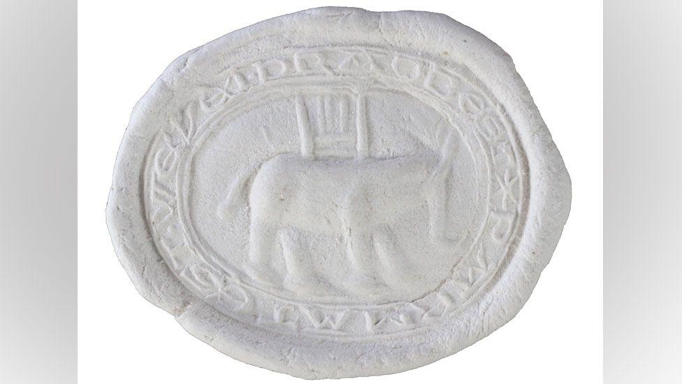 An impression in white wax of an oval seal matrix. It shows a narrow band on its circumference which is inscribed with letters. Its middle shows a portly-looking elephant with broad legs and tail and a trunk. On its back is a castle or howdah.