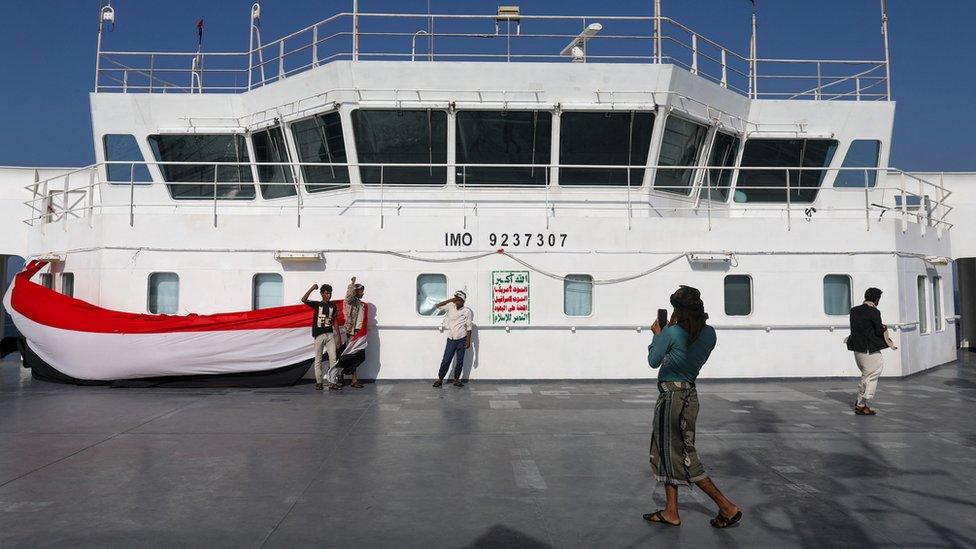 A man takes a picture of a group of people unfurling the Yemeni flag onboard the Galaxy Leader while the Houthi flag has ben painted on a section of the ship in the centre of the image.