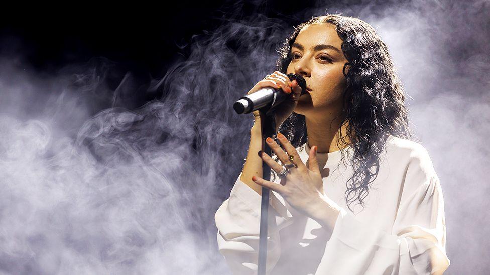 Charli XCX, a female singer performing on stage, singing into a black microphone while wearing a white outfit, while smoke effects surround her.