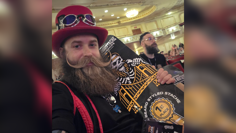 A man - Lewis Aylmer - wearing a red top hat with blue-tinted glasses on and a black and red outfit. He is holding a black and yellow skateboard with a fist place ribbon on. People can be seen in the background. He is in a theatre-like room.