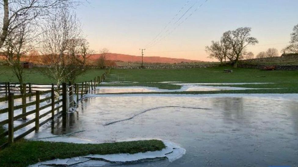 Fields in Airton, North Yorkshire, covered in frozen flood water. Sunrise can be seen in the distance. 