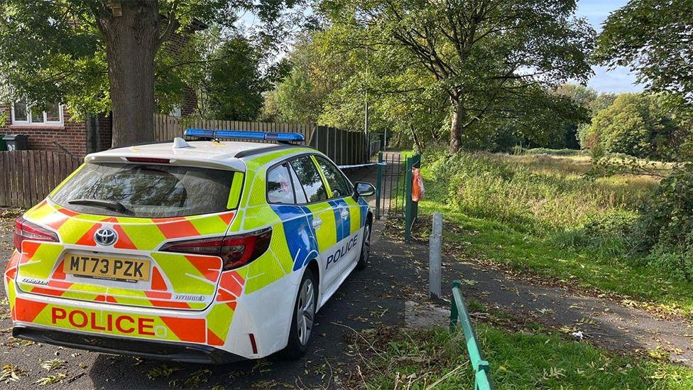 Police car parked near the scene. It is by a pathway fenced off between houses and a green park area