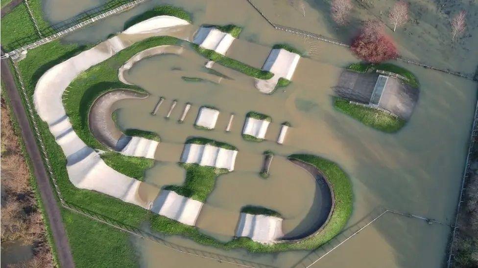 An aerial view of a flooded BMX track where ramps and hills pop out of a large surface of brown water