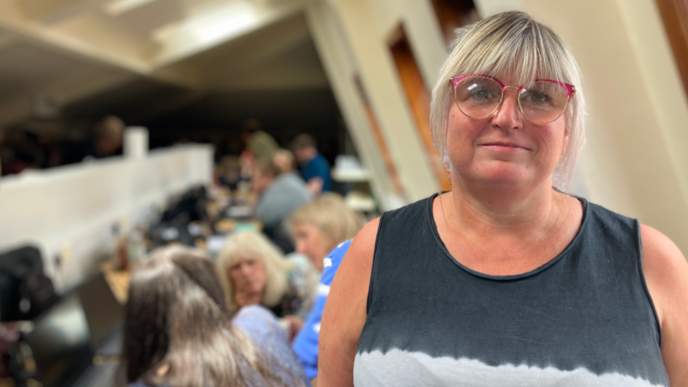 A woman with blonde hair and red-rimmed glasses, and wearing a sleeveless black and white shirt, stands in an office and smiles. People can be seen talking and walking behind her.