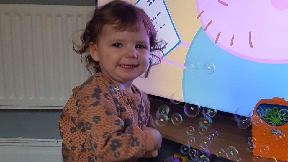 Maya, a young girl stands in front of a TV playing Peppa Pig and next to a bubble machine