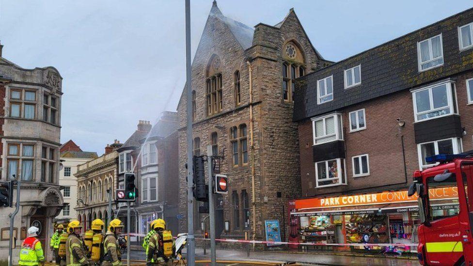 A group of firefighters standing next to a red and white cordon, with a smoking building behind it.