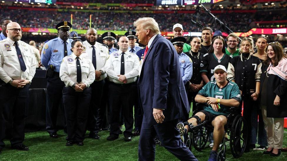 Donald Trump walks from right to left on the pitch at the super bowl, behind him are people watching him